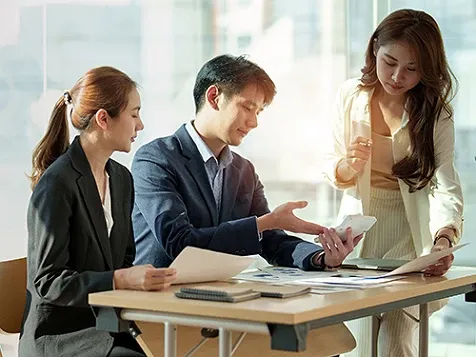 A man and a woman seated at a desk, while another woman is standing and all three are looking at documents